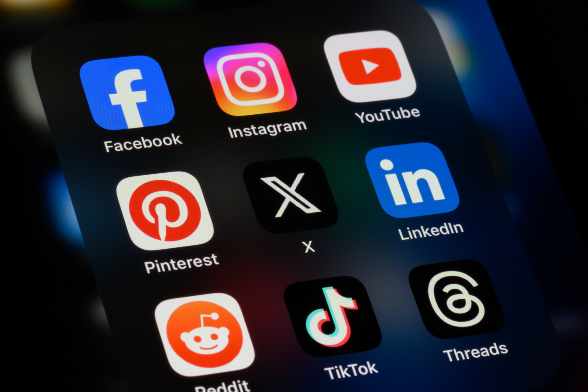 A smartphone screen displaying a folder of popular social media app icons, including Facebook, Instagram, YouTube, Pinterest, X, LinkedIn, Reddit, TikTok, and Threads.