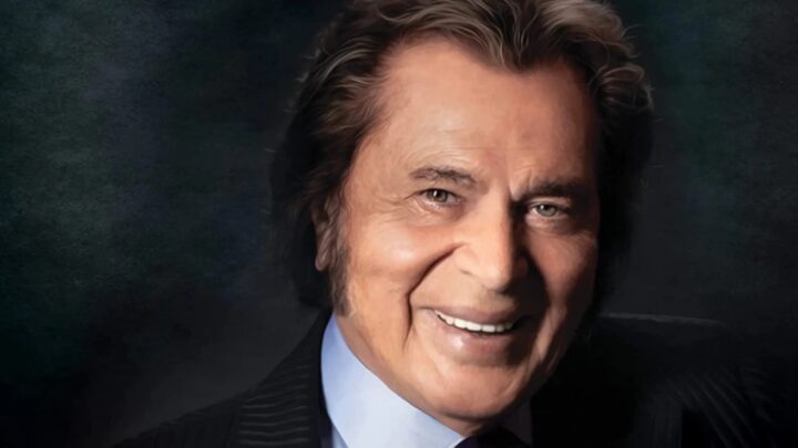Portrait of singer Engelbert Humperdinck smiling in a dark suit against a softly lit studio background.